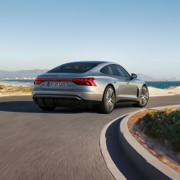 Ein silberner Audi S e-tron GT fährt auf einer kurvigen Küstenstraße mit Blick auf das Meer und eine Stadt im Hintergrund.