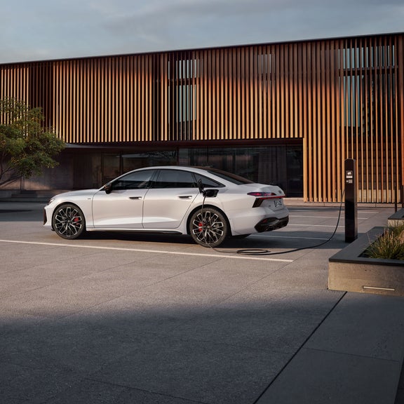 Die A6 Limousine e-hybrid in weiß steht seitlich vor einer Gebäudefassade und ist dabei an eine Ladestation angeschlossen.