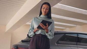 Eine lächelnde Frau steht unter einem Carport und bedient ein Tablet. Im Hintergrund steht ein silberfarbener Audi A6 e-tron.