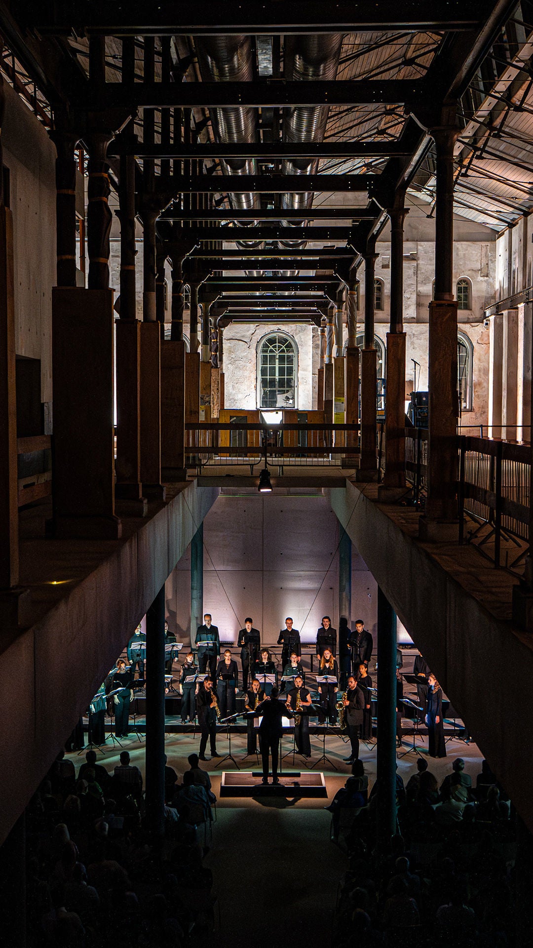 Orchester mit Dirigent spielt in einer großen Industriehalle; Publikum sitzt davor, dramatische Beleuchtung, dunkle Stahlträger.