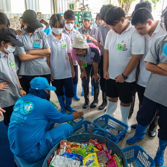 Menschen stehen im Kreis. Vor ihnen hockt ein Mann und zeigt ihnen Dinge.
