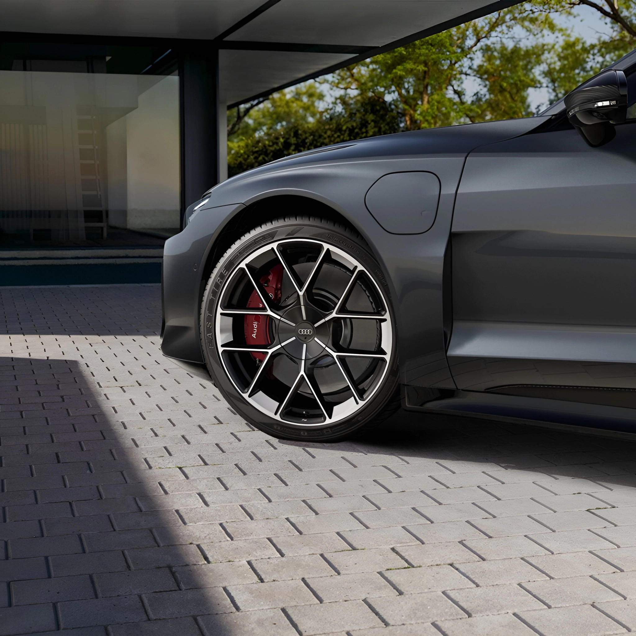 Dark grey Audi car on grey brick driveway, showing front wheel with red brake caliper and black rim with silver accents.