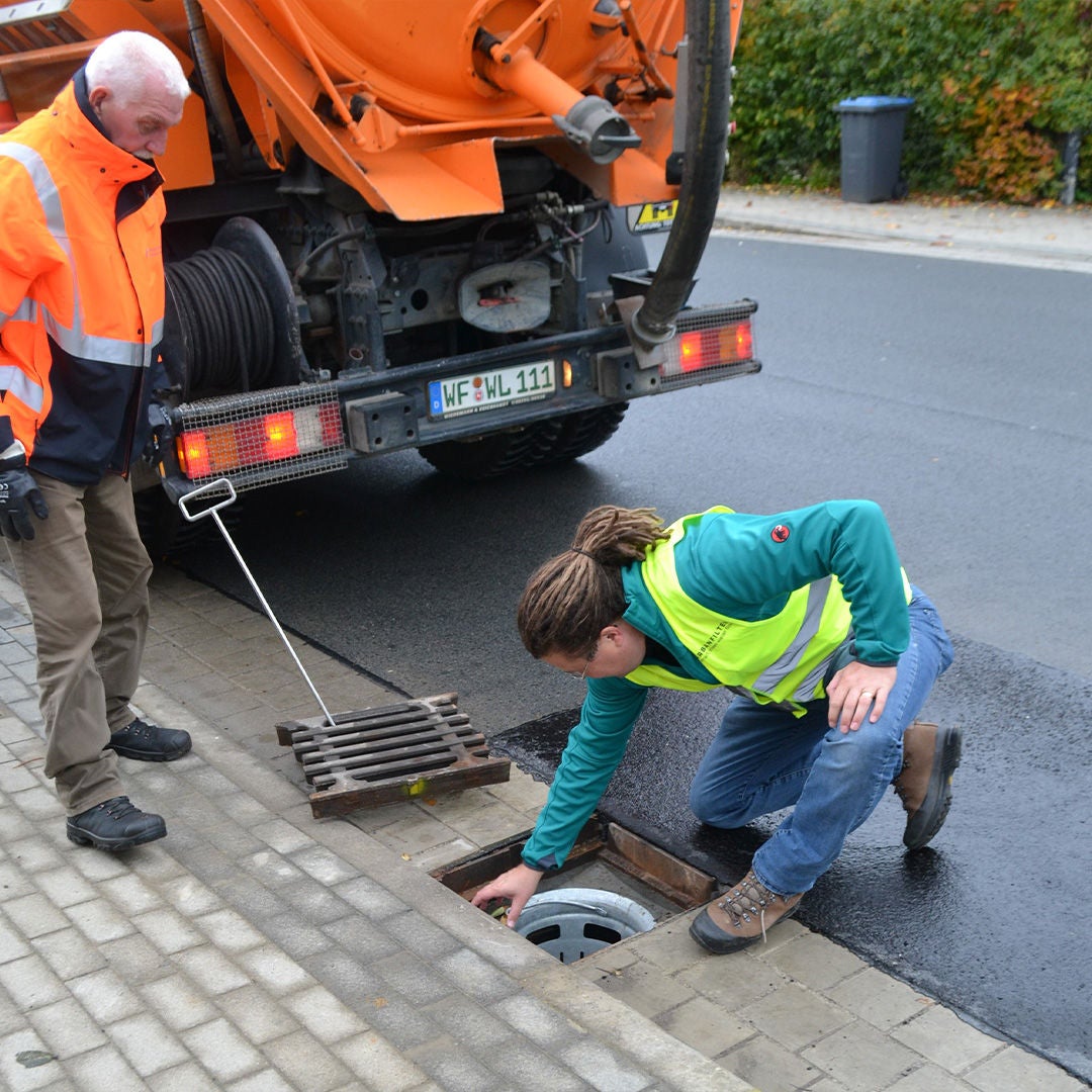 Two men at an open manhole cover, one kneeling and inspecting the opening, an orange truck is parked next to them.  