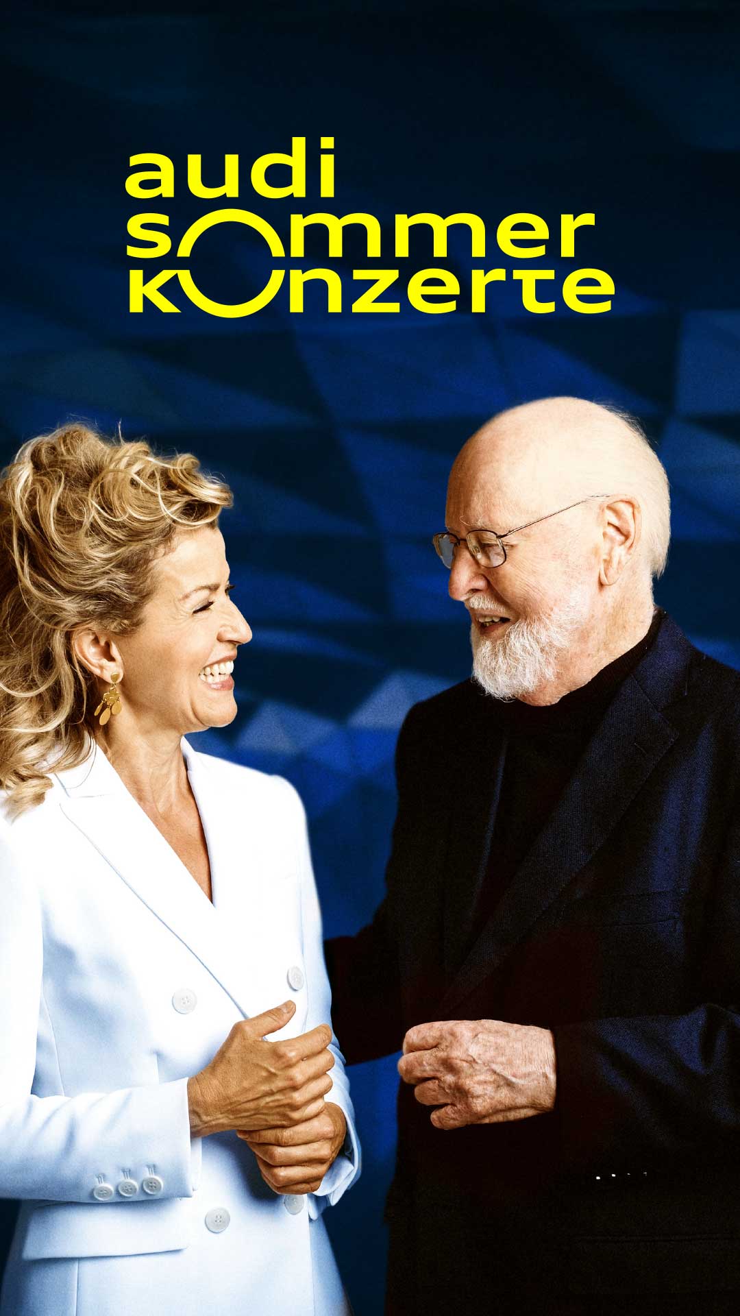 Ältere Dame und älterer Herr schauen sich an und lächeln, im Hintergrund blau und das gelbe Logo der Audi Sommerkonzerte.