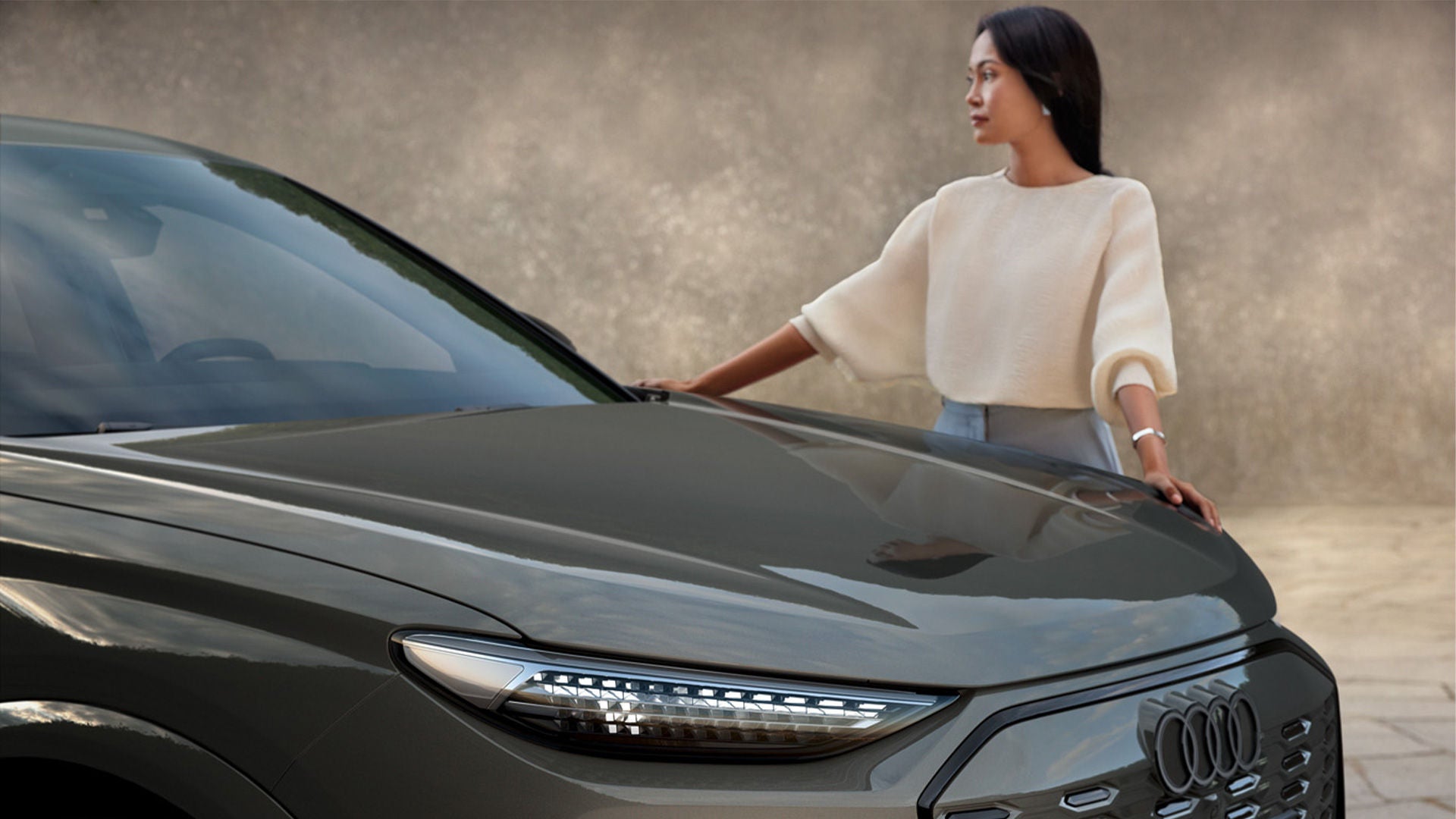 Eine asiatische, junge Frau steht neben der Front eines grauen Audi Q6 Sportback e-tron mit schmalem Scheinwerfer und schwarzen Audi Ringen.