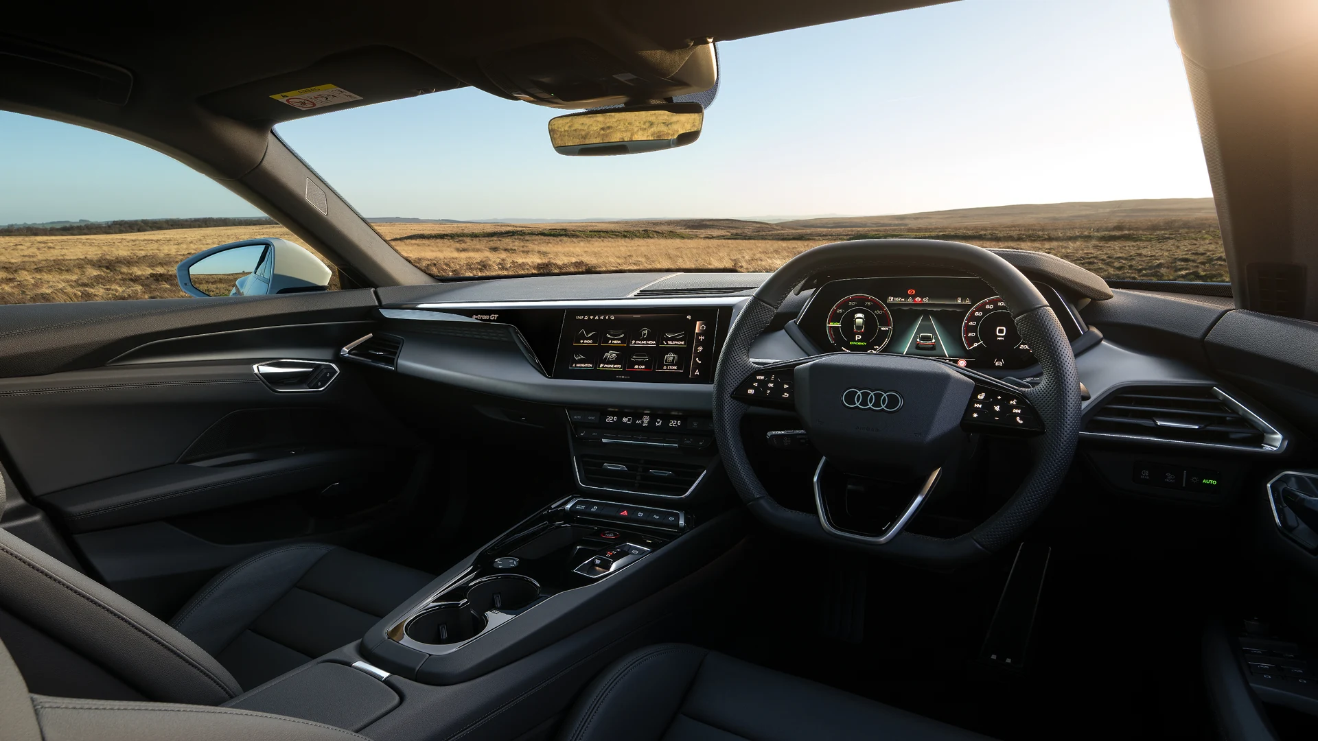 Audi e-tron GT interior viewed from the driver's seat, featuring a multifunction steering wheel, digital instrument cluster and central touchscreen display. The premium cabin includes black leather seats, a streamlined centre console with cup holders and integrated controls. Open countryside with rolling fields is visible through the windscreen under bright daylight.