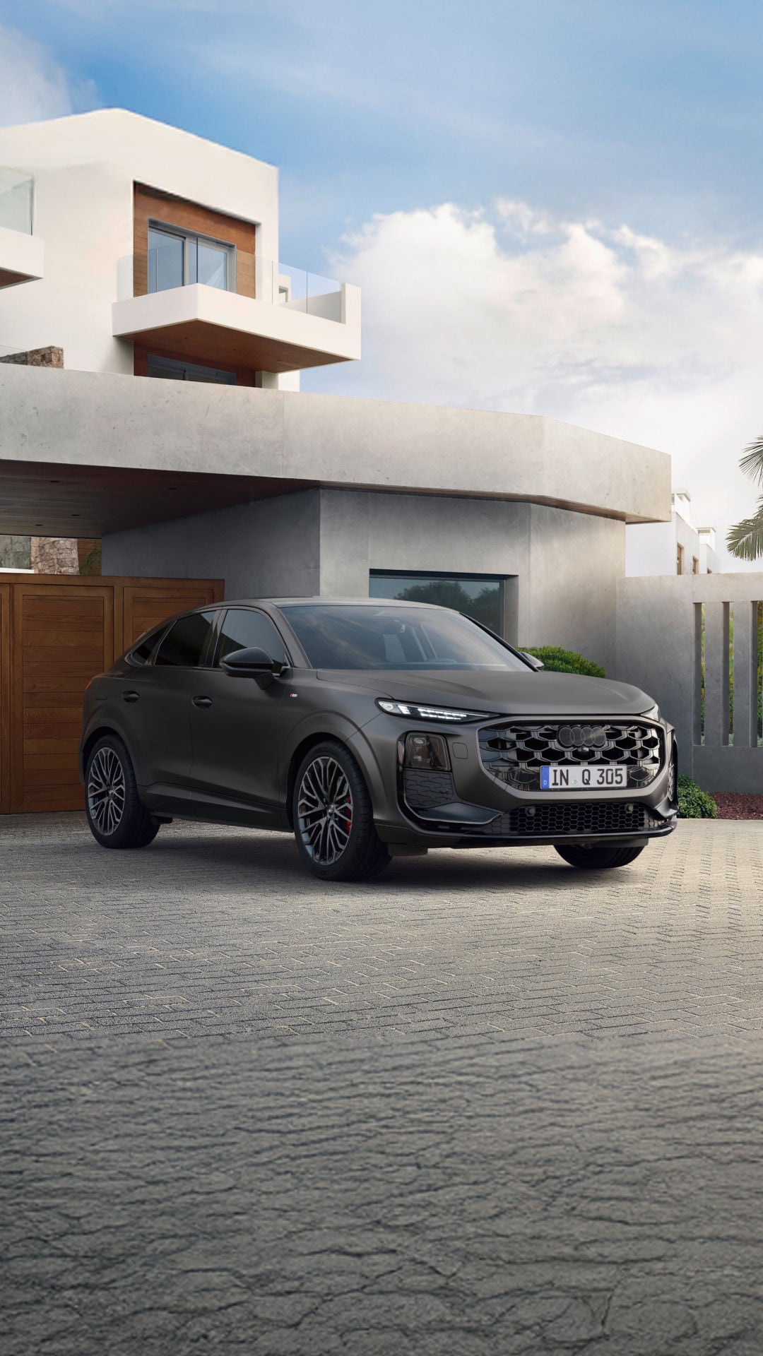 A matte black Audi Q3 parked in front of a modern house with palm trees and wooden garage doors in daylight.