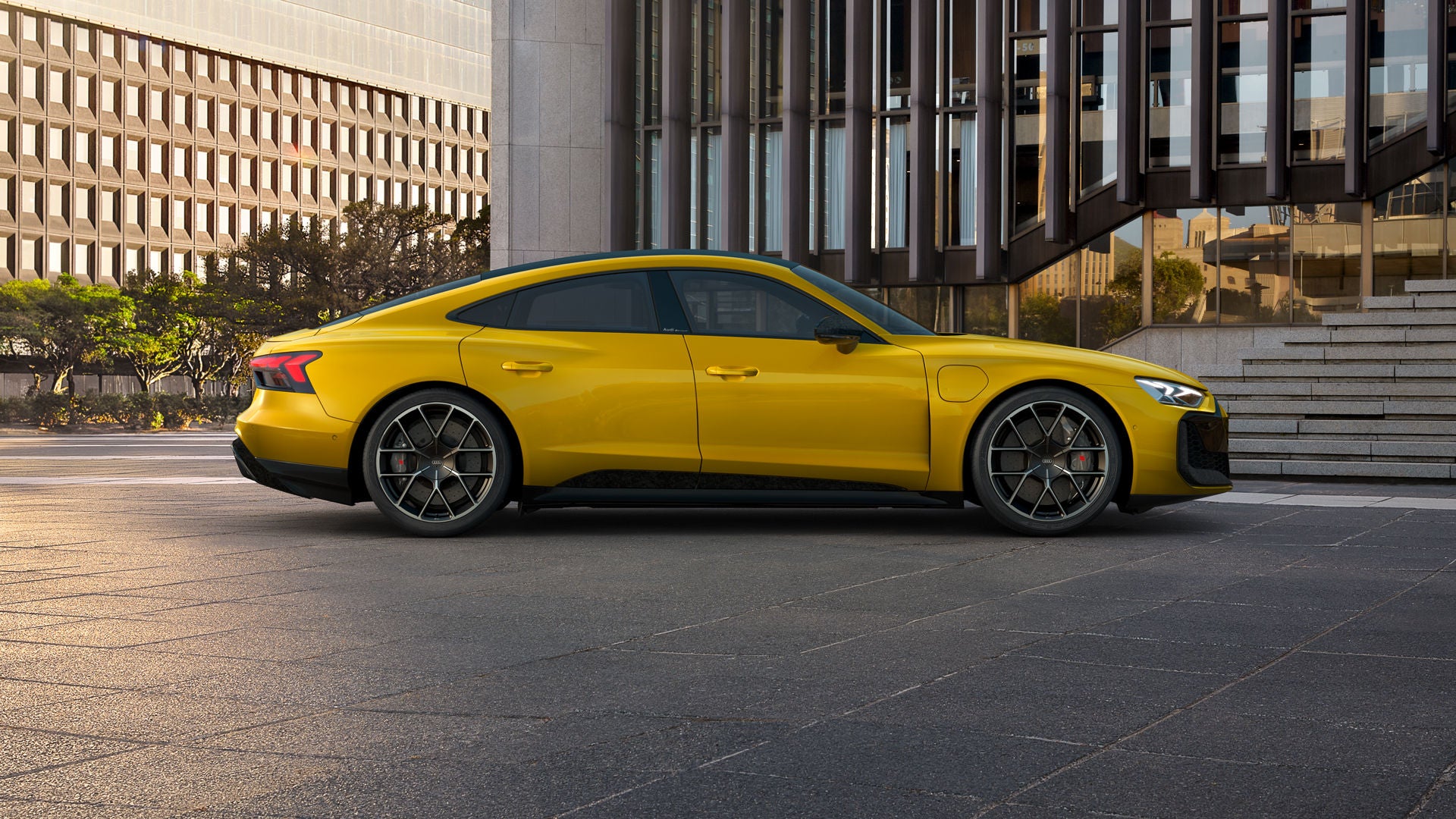 Gelber Audi RS e-tron GT performance geparkt vor einem modernen Gebäude mit Glasfassade und Treppe bei Sonnenuntergang.