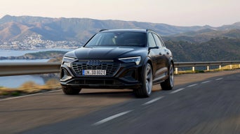 A black Audi vehicle driving on a coastal road with mountains and a bay in the background