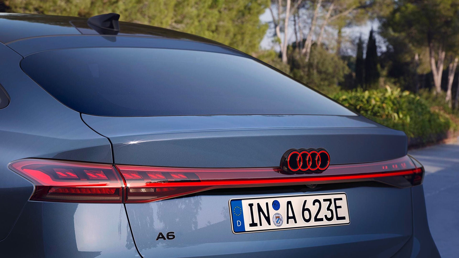 Close-up of a blue Audi A6's sleek rear, featuring distinctive red taillights and chrome Audi rings. Sunny day with blurred greenery in the background.