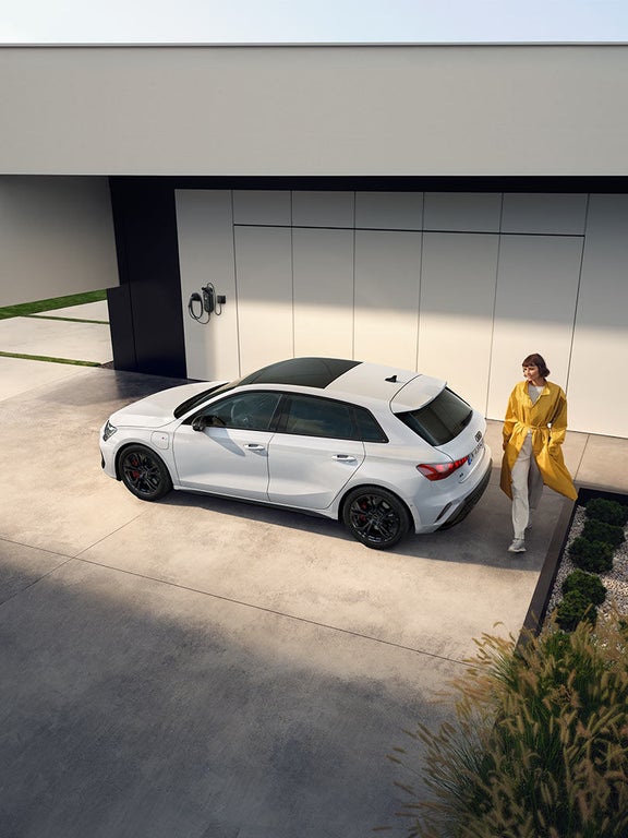 Schräg von oben: Frau in gelbem Mantel steht neben weißem Audi A3 Sportback TFSI e auf heller Betonfläche.