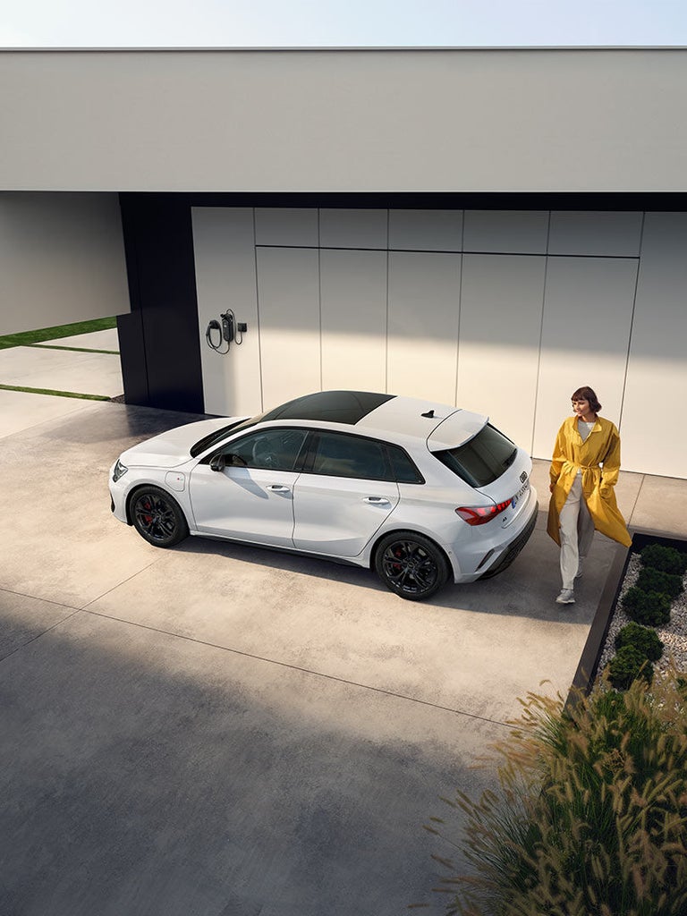 Schräg von oben: Frau in gelbem Mantel steht neben weißem Audi A3 Sportback TFSI e auf heller Betonfläche.