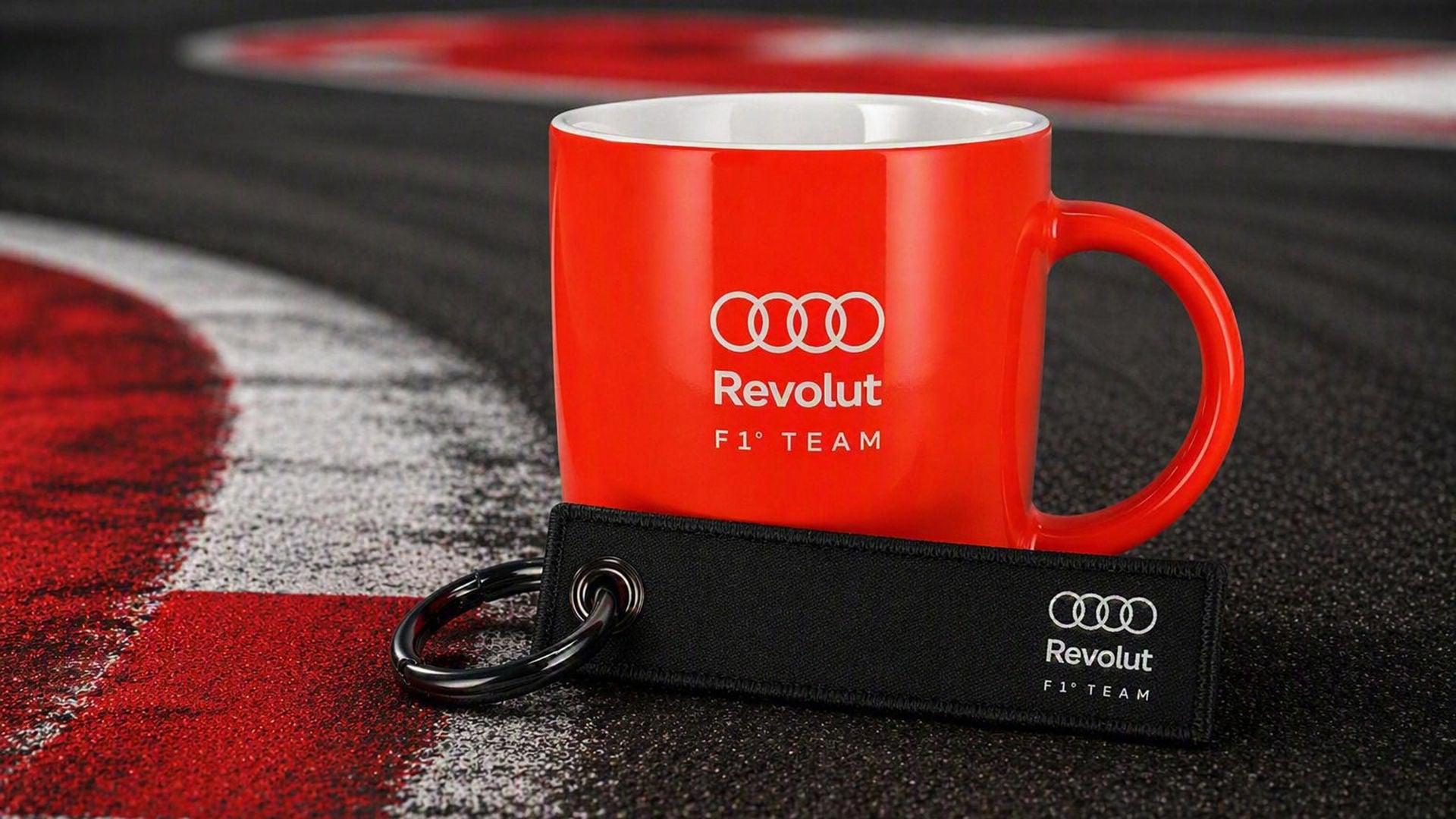 Eine rote Tasse mit Audi und Revolut Logo und ein Schlüsselanhänger liegen auf einer Rennstrecke auf dem Boden