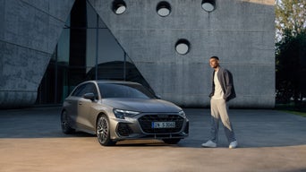 Ein Mann steht neben einem grauen Audi S3 vor einem modernen Betongebäude mit runden Fenstern bei Sonnenuntergang.