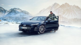 Blauer Audi steht vor verschneitem Bergpanorama. Mann steigt in das Auto ein.