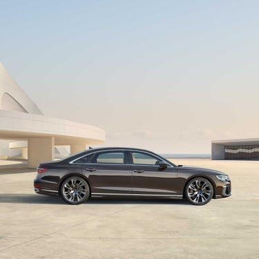 Seitenansicht eines braunen Audi A8 L auf hellem Platz vor modernem, weißem Gebäude.
