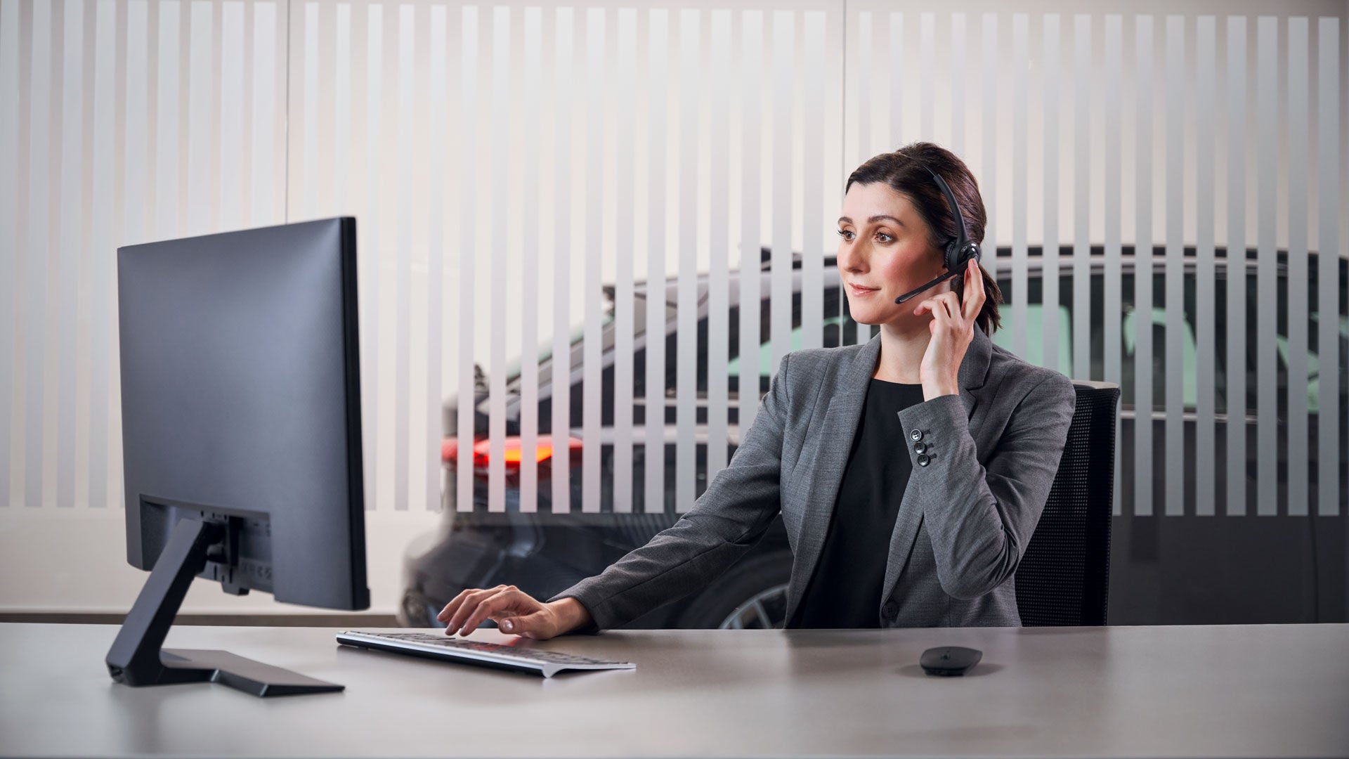 Mitarbeiterin mit Headset sitzt am Schreibtisch vor einem Monitor in einem Büro mit Auto im Hintergrund.