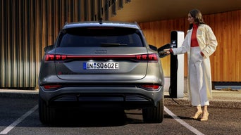 Der SQ6 SUV e-tron steht auf einem Parkplatz vor einer Gebäudefassade und wird von einer jungen Frau an eine Ladestation angeschlossen.