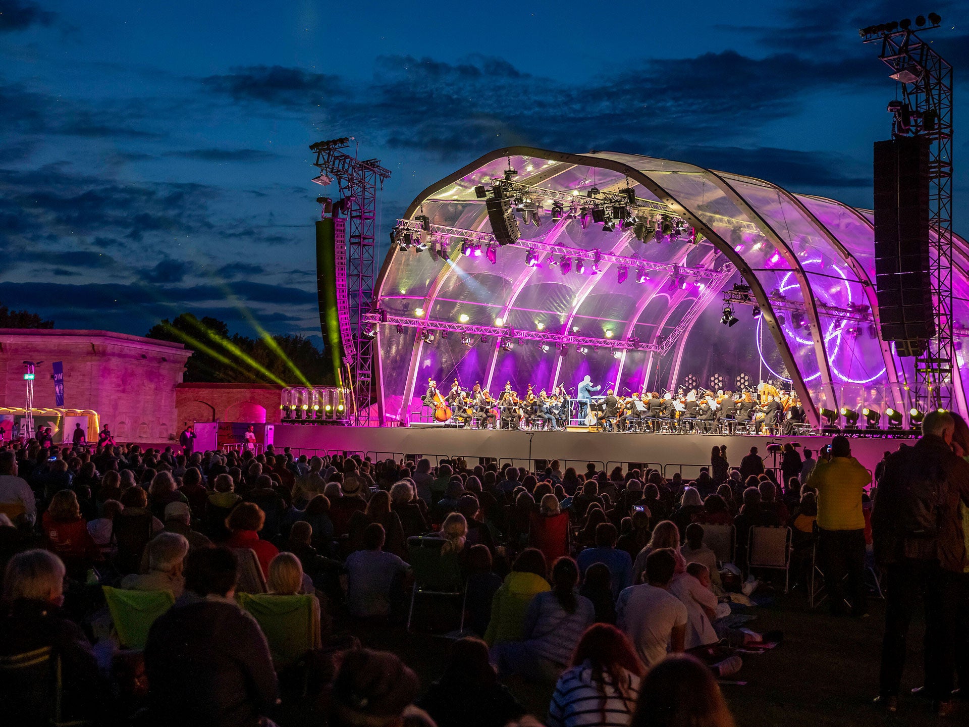 Ein Orchester auf einer bunt beleuchtete Open Air Bühne unter einer Glaskuppel. Davor steht das Publikum.