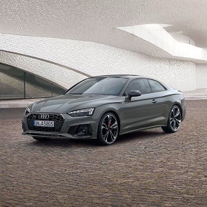 Grey Audi on cobblestone, against modern architecture with wavy, textured walls. The car's design exudes luxury and sophistication.