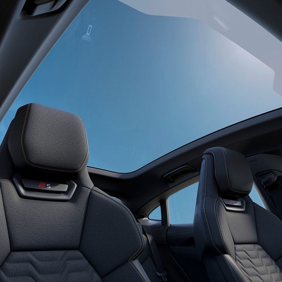 Interior view of a modern car featuring sporty seats with "S" badge and a panoramic sunroof showcasing a clear blue sky.