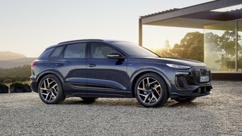 Der Q6 SUV e-tron steht seitlich auf der Kiesfläche vor einem modernen Haus und wird dabei von der untergehenden Sonne angestrahlt.