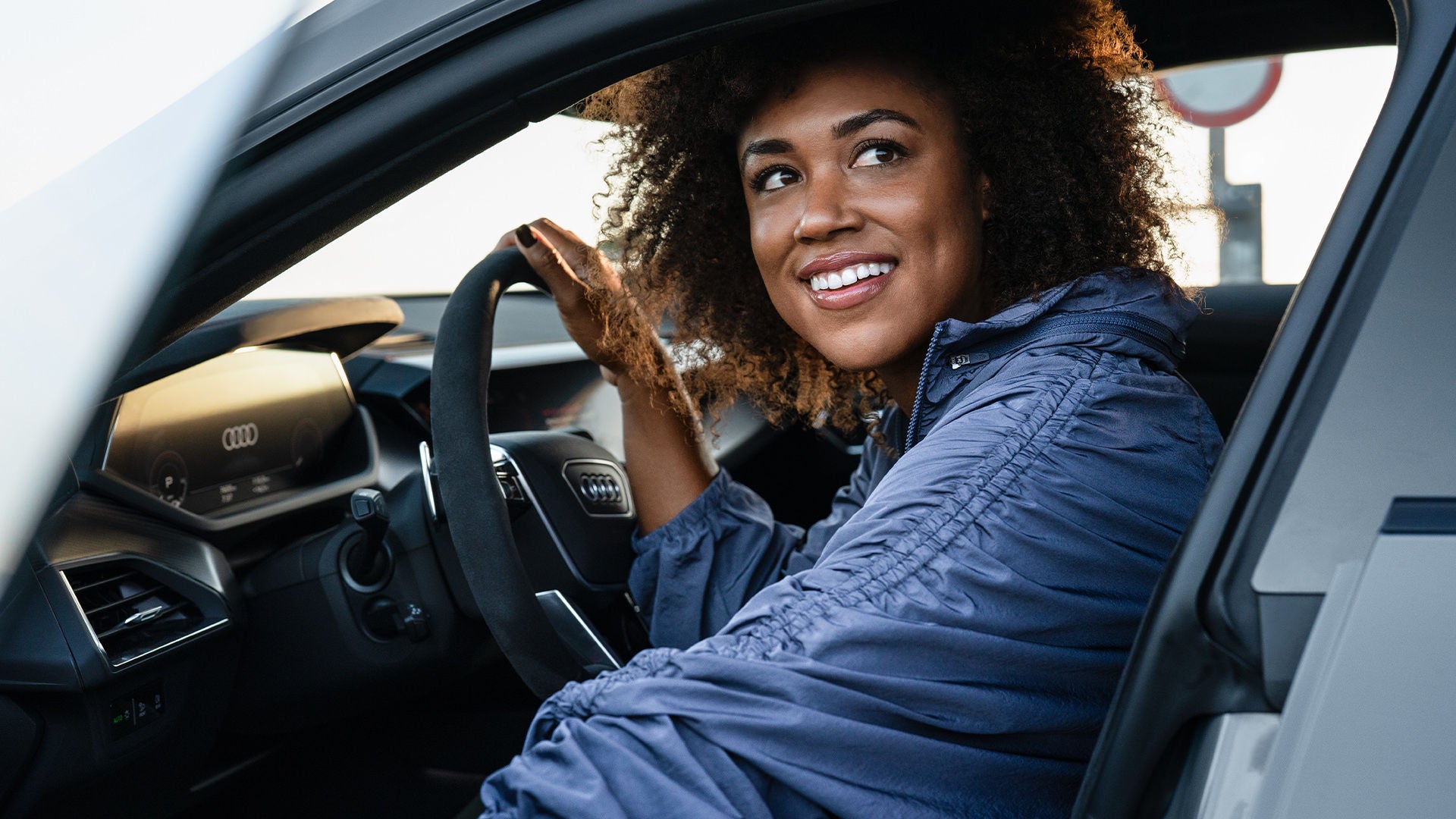 Frau mit lockigem Haar sitzt lächelnd am Steuer eines Autos und schaut zur Seite.  