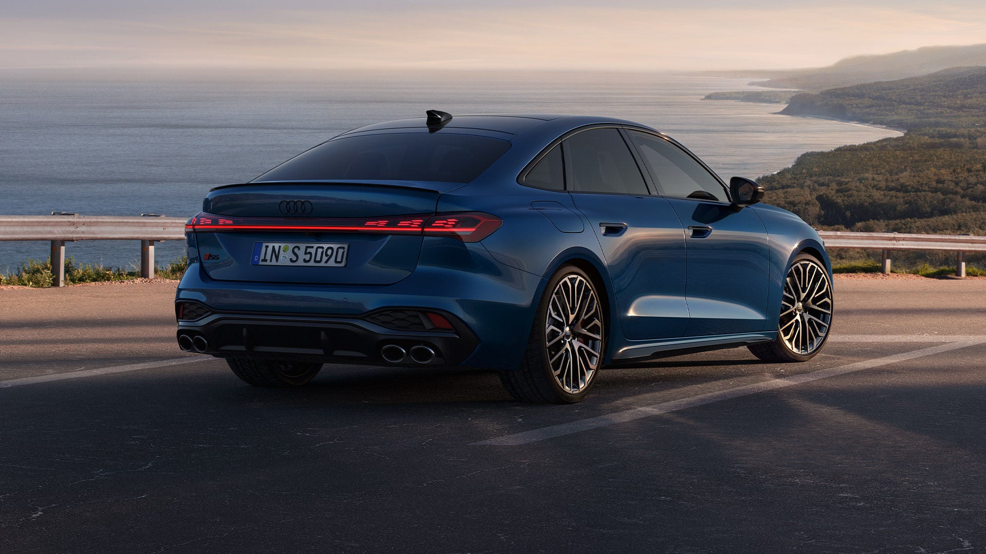 Blaue Audi S5 Limousine steht auf Küstenstraße mit Blick auf das Meer und bewaldete Hügel im Hintergrund.