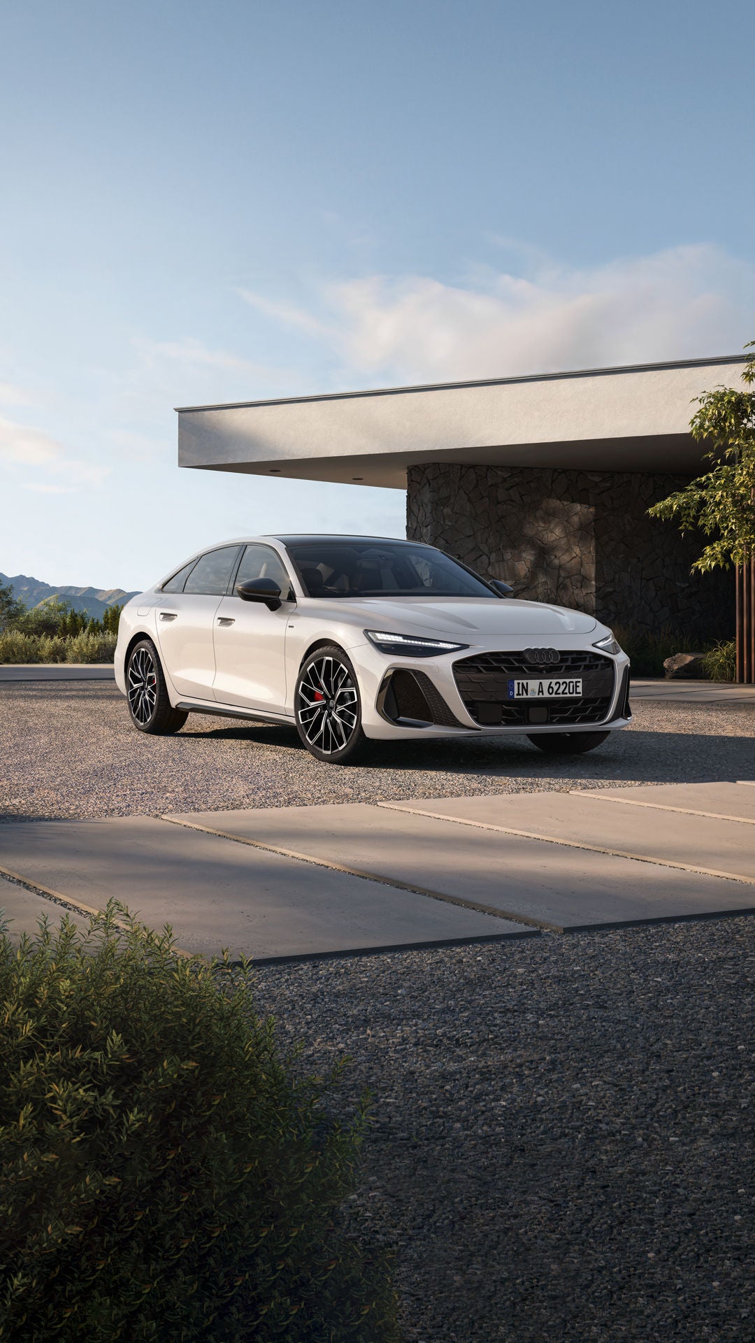 Die weiße A6 Limousine e-hybrid aus schräger Frontansicht parkt vor einem modernen Steinhaus.