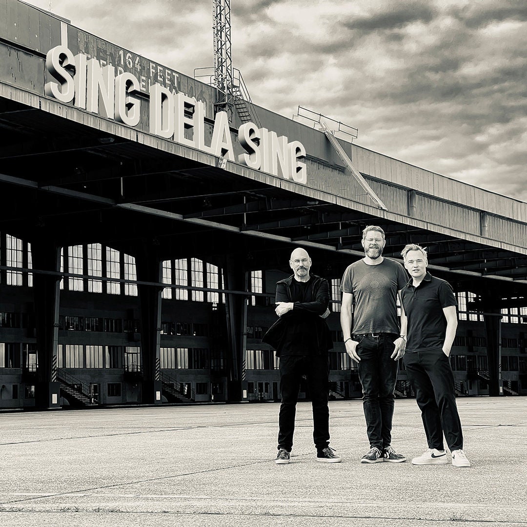Drei Männer stehen vor einem Industriegebäude mit dem Schriftzug „Sing dela Sing", Schwarzweißfoto.
