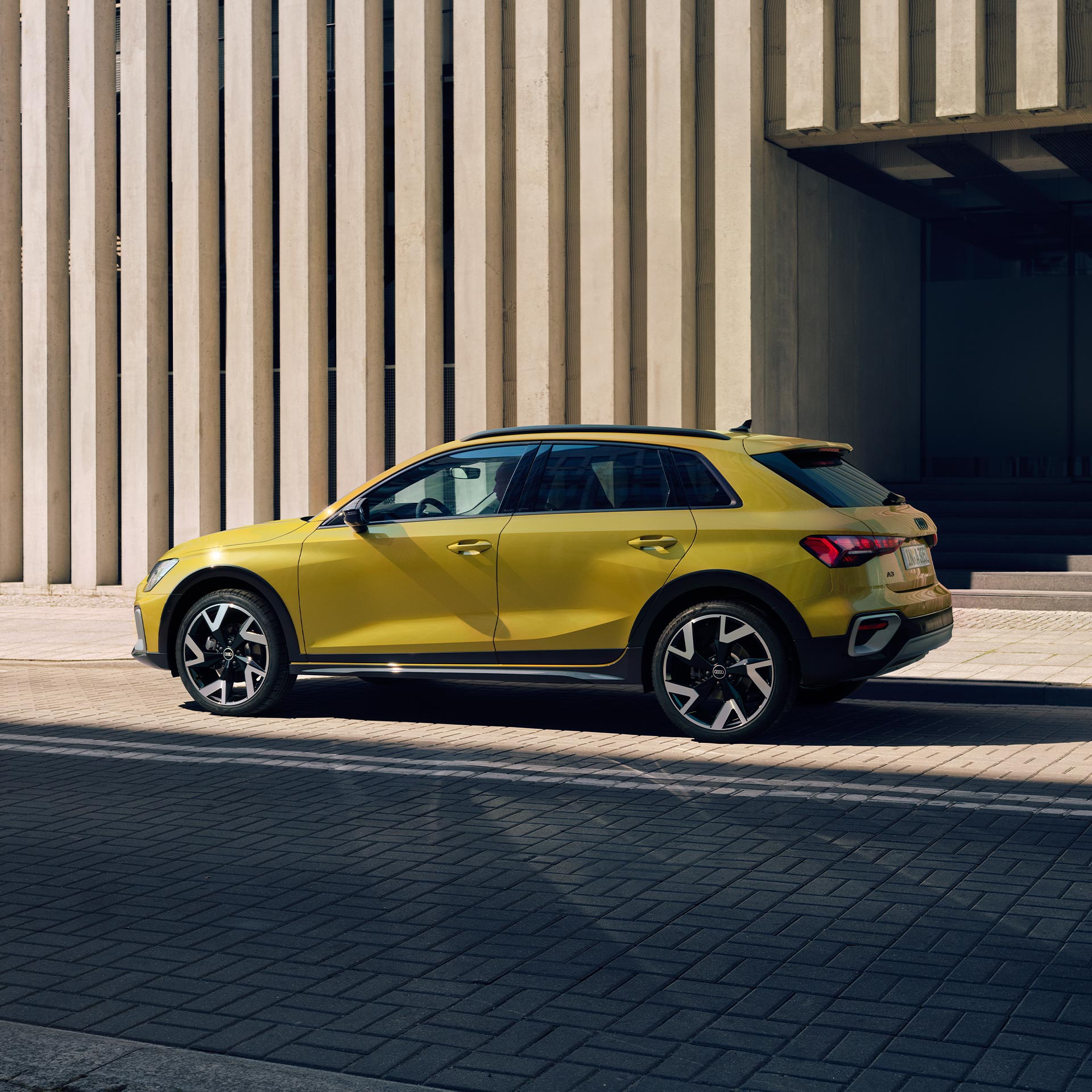 Gelber Audi A3 allstreet seitlich vor einem modernen Gebäude mit vertikalen Betonstreben fotografiert.