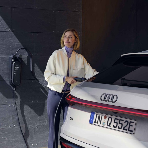En ung kvinna ansluter Q5 Sportback e-hybrid till laddstationen som är monterad på husets vägg. 