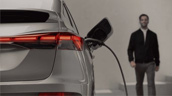 A sleek silver electric vehicle is plugged in and charging, with a blurred figure walking in the background.