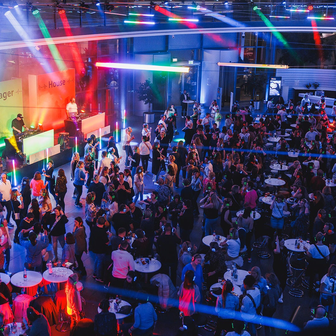 Eine Menschenmenge tanzt in einem Veranstaltungsraum mit Kopfhörern, vorne steht ein DJ an einem Pult und es leuten bunte Lichter