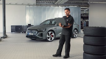 In einer Werkstatt hält ein Mechaniker in schwarzer Audi Arbeitskleidung ein Laptop, grauer Audi SUV steht im Hintergrund, Reifenstapel im Vordergrund.