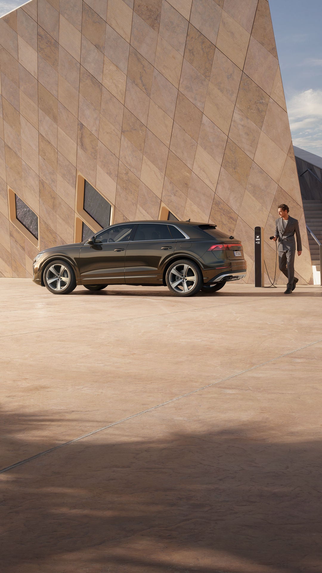 De Q8 e-hybride staat geparkeerd in een schuin zijaanzicht voor een futuristisch gebouw.  Achter het voertuig staat een modieus geklede jongeman, met de laadstekker van een laadstation in zijn hand.