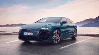 Ein blauer Audi steht auf nasser Straße am Wasser, Berge und heller Himmel im Hintergrund.