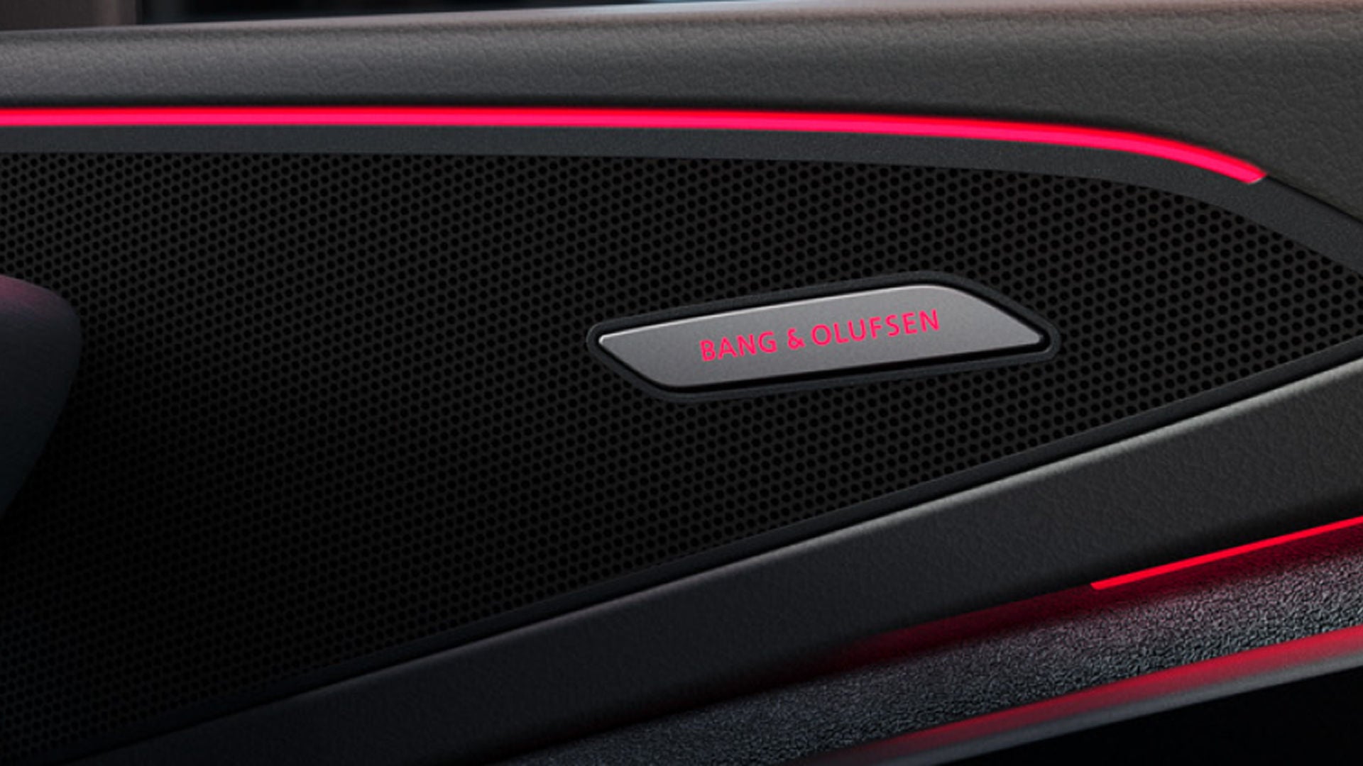 Close-up of a car speaker with a black perforated mesh, featuring a silver "Bang & Olufsen" logo. Red ambient light highlights the sleek design.