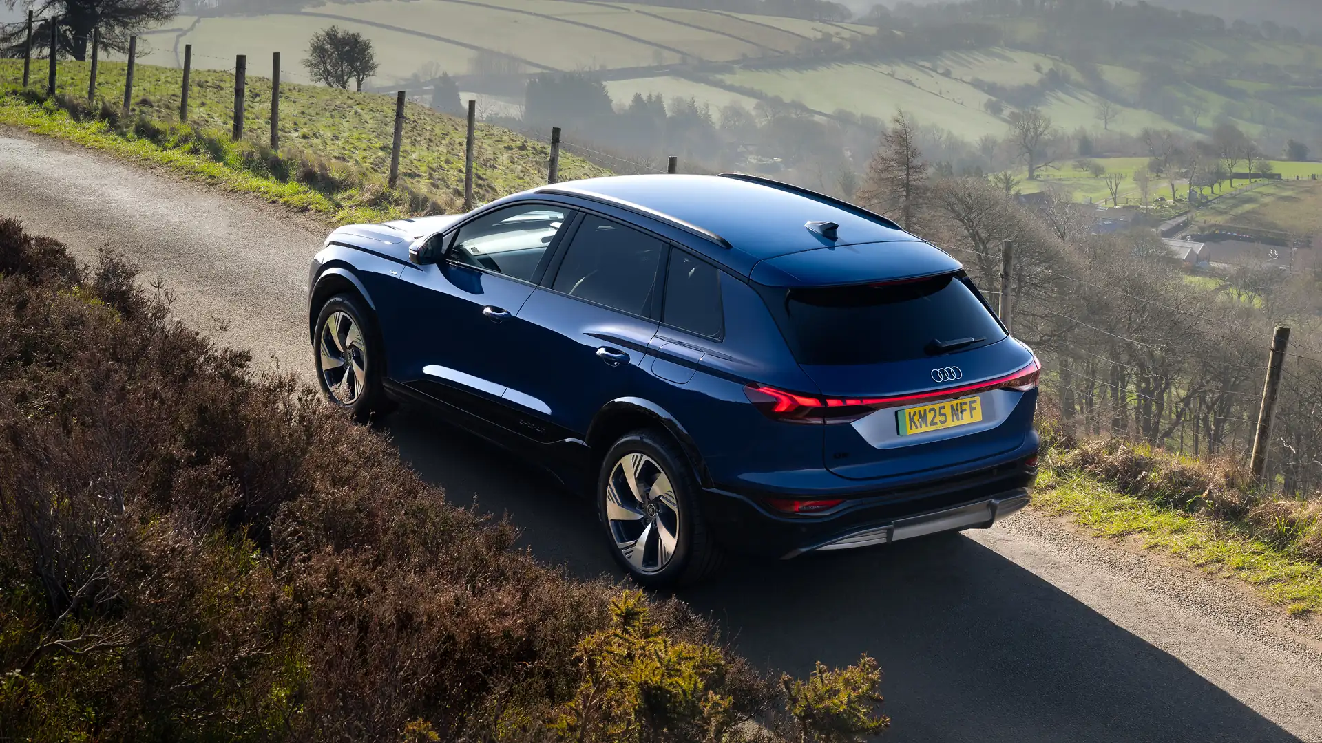Blue Audi Q6 e-tron viewed from the rear three-quarter angle on a winding rural road. The SUV features a full-width rear LED light bar, aerodynamic alloy wheels and a sculpted body design. Rolling green hills, fence posts and patchwork countryside fields stretch across the background, while bright daylight highlights the vehicle's contours and surrounding landscape.