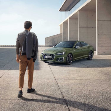 Man in a jacket looking at a green Audi on a sunny rooftop parking.