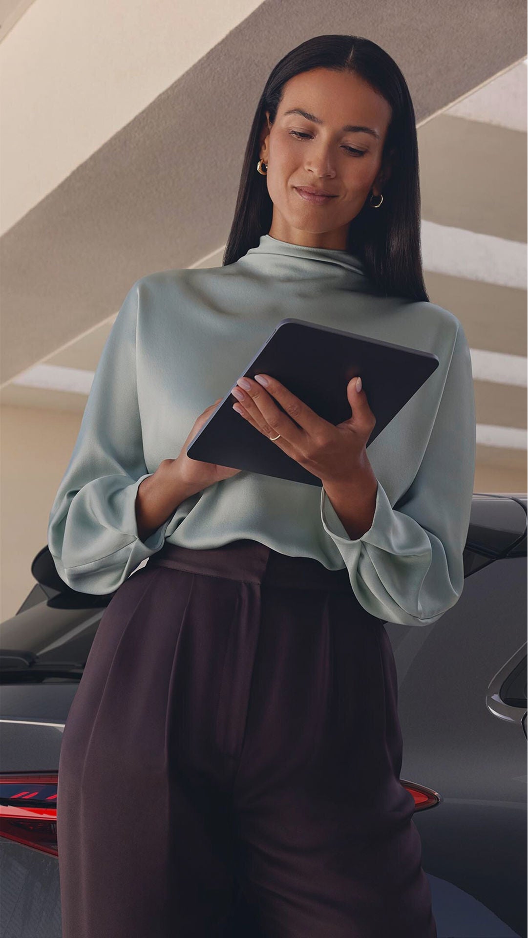 A person holding a tablet stands in front of a car, under a modern, sunlit covered structure.