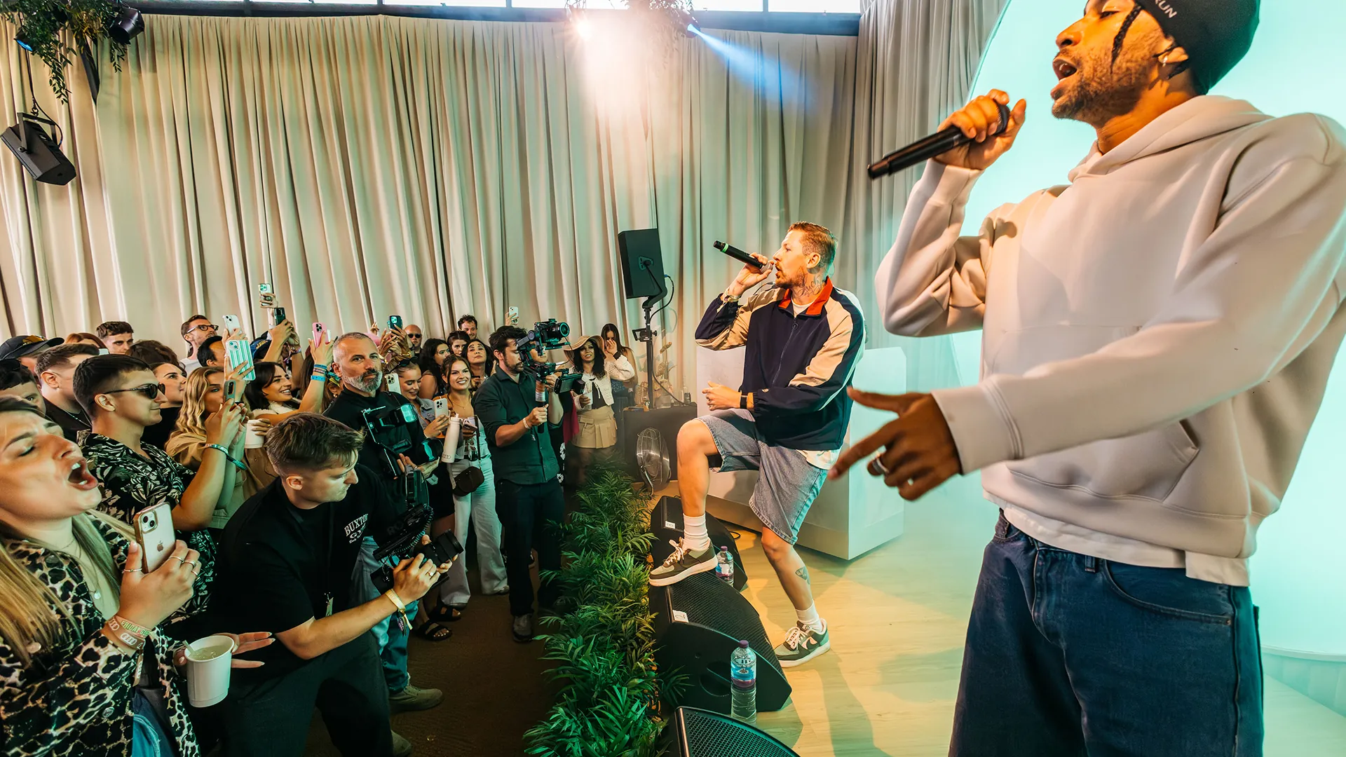 Two performers sing into microphones on stage as a crowd films and cheers, lit by warm stage lighting under a curtained backdrop. Two performers sing into microphones on stage as a crowd films and cheers, lit by warm stage lighting under a curtained backdrop.