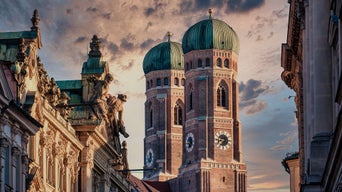 Zwei Türme der Frauenkirche in München mit grünen Zwiebeldächern zwischen historischen Fassaden bei Abendhimmel.