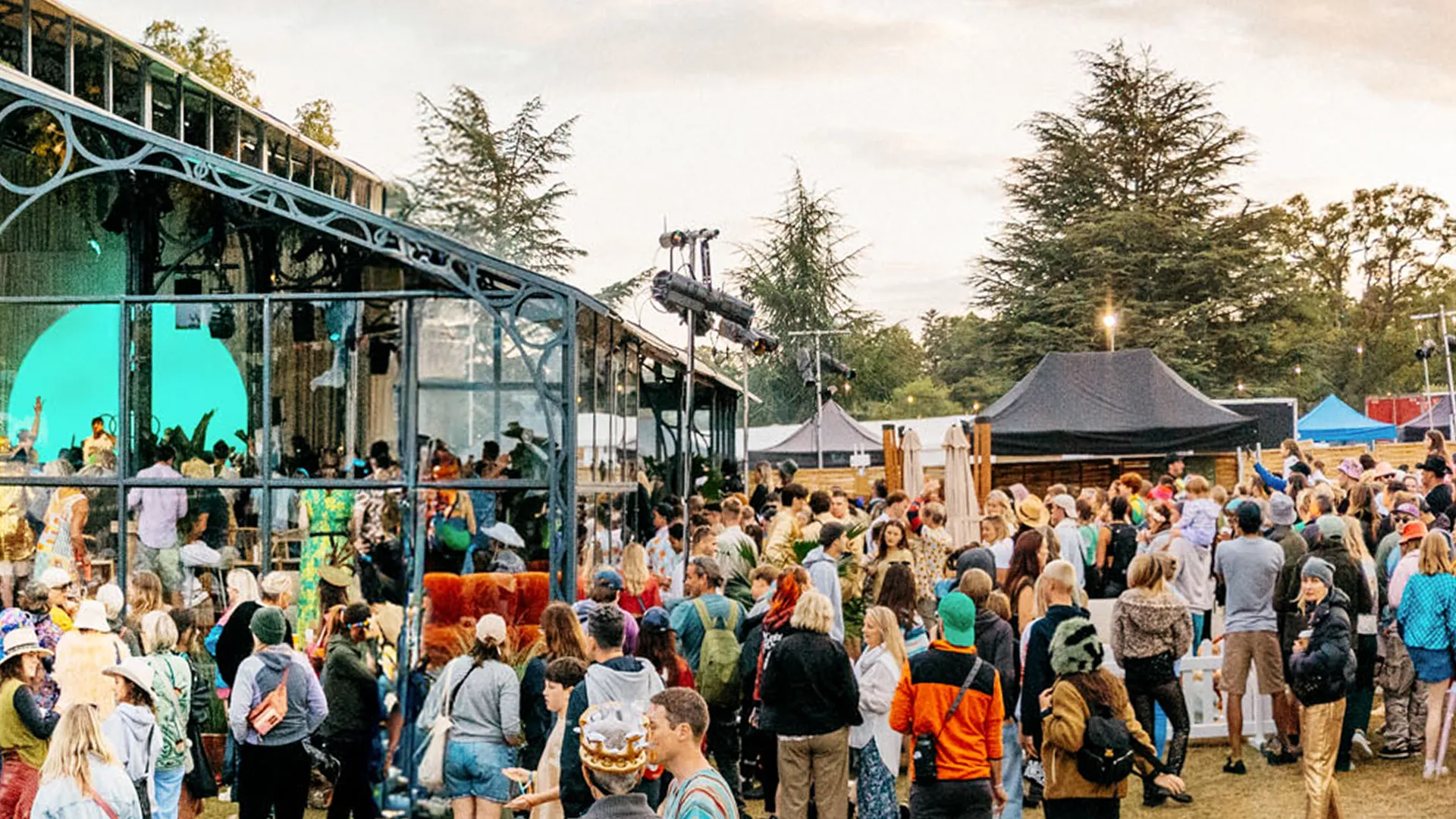 A crowd gathers outside a glass-walled pavilion at dusk, with stage lighting visible inside and festival tents in the background. A crowd gathers outside a glass-walled pavilion at dusk, with stage lighting visible inside and festival tents in the background.
