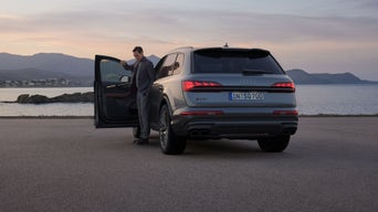 Ein Mann steigt aus einem silbernen Audi SQ7 SUV, der am Wasser vor einer Bergkulisse geparkt ist.