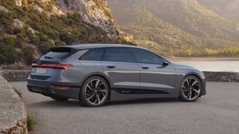 Der A6 Avant e-tron steht seitlich auf einem Kiesparkplatz vor einem See. Im Hintergrund sind von Bäumen bewachsene Berge zu sehen.