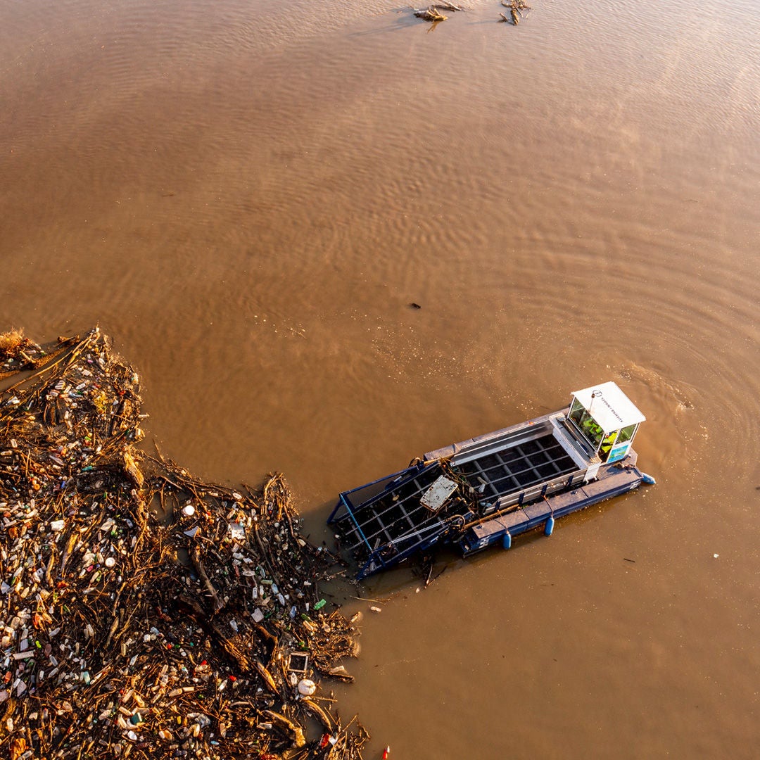 Bird's eye view of a river with brown-coloured water. A rubbish collection boat picks up rubbish from the water.