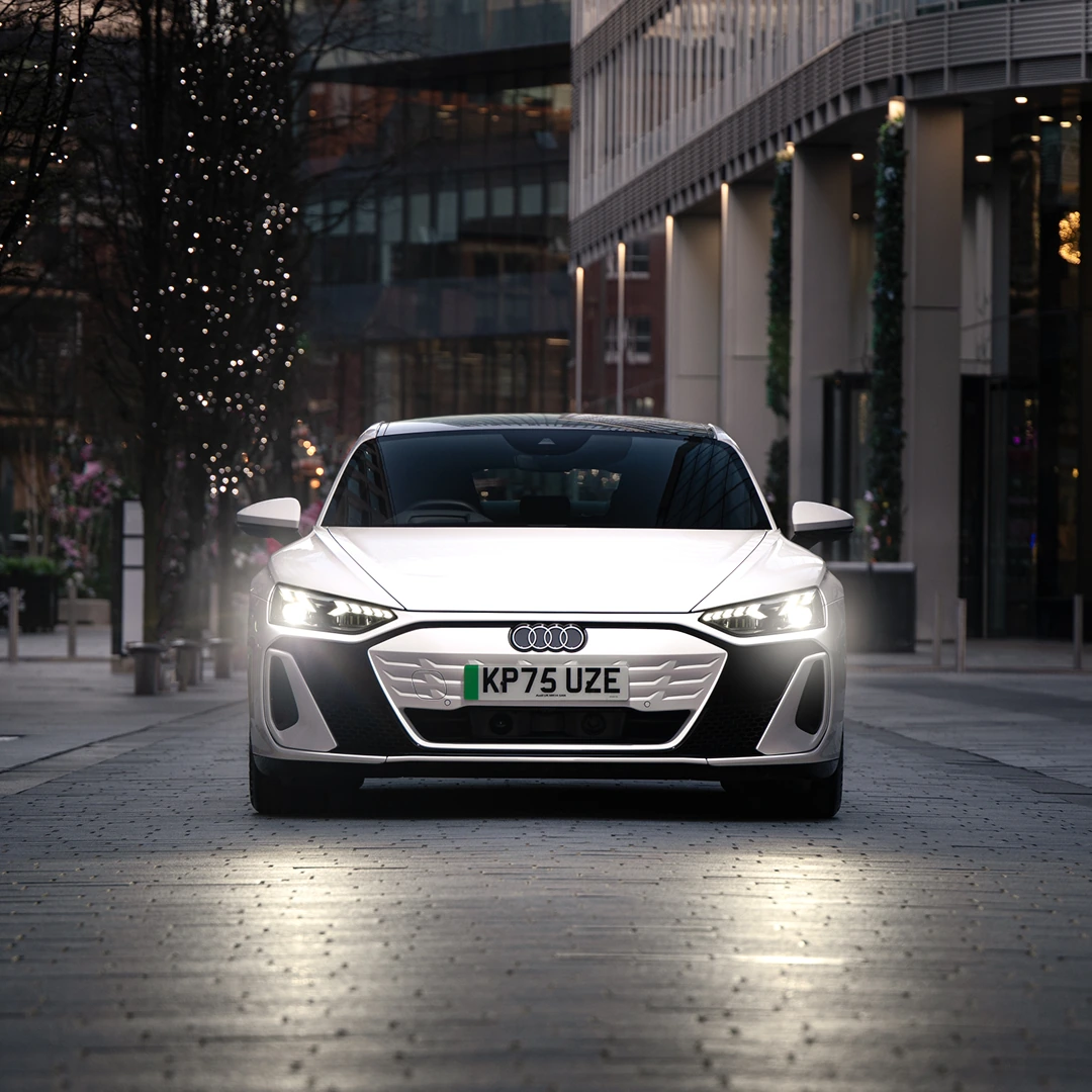 White Audi e-tron Sportback viewed from the front on a city street at dusk. The vehicle's LED headlights are illuminated, highlighting the sculpted front fascia and aerodynamic design. Modern office buildings and trees decorated with string lights line the street, while reflections from the headlights are visible on the paved road surface.