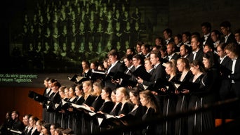 Großer Chor in schwarzer Konzertkleidung singt auf einer Bühne mit projiziertem Text im Hintergrund.