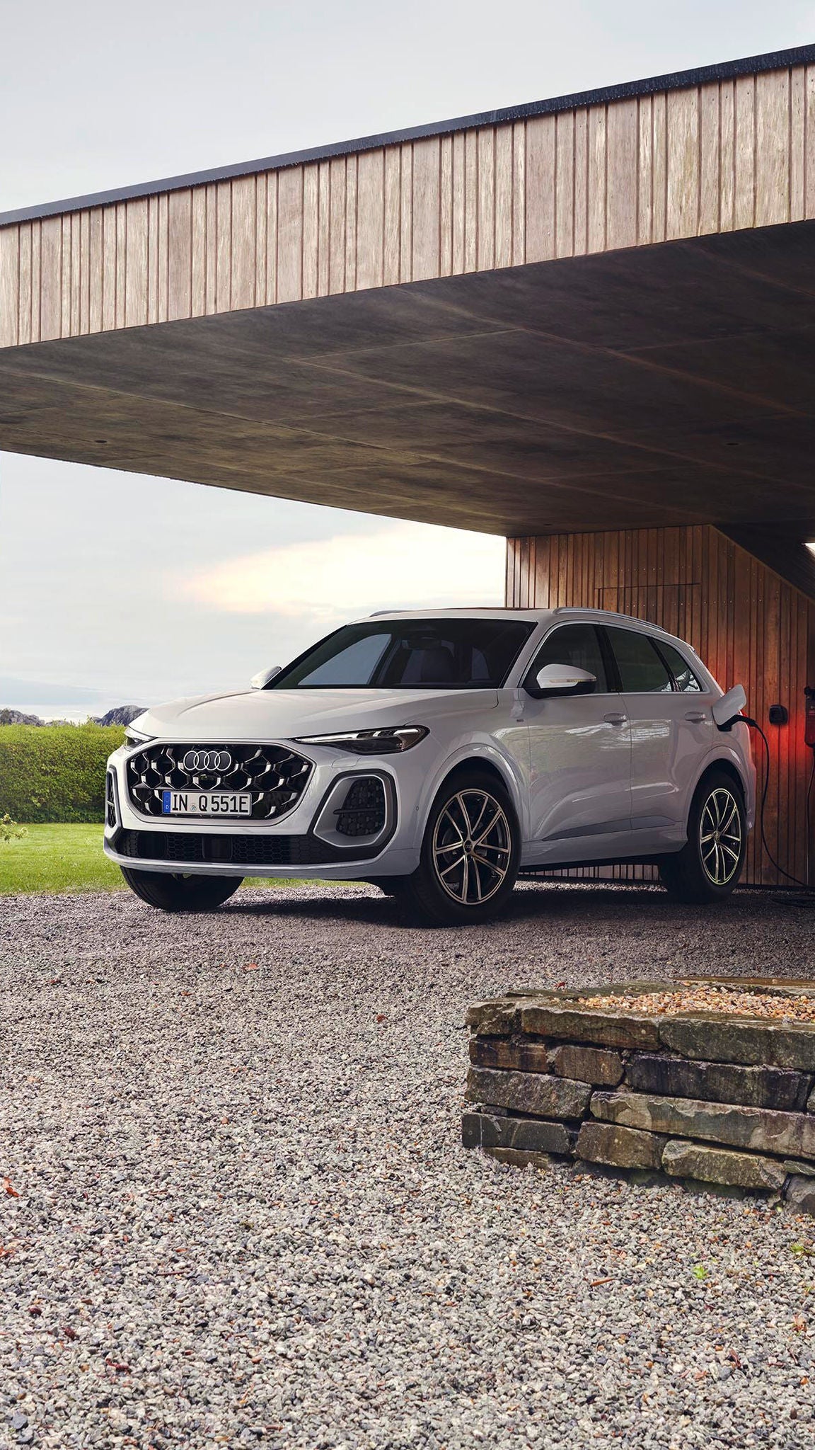 Audi Q5 e-hybrid parkerad i ett öppet garage på en grusuppfart, till höger går en person och håller i ett paraply.